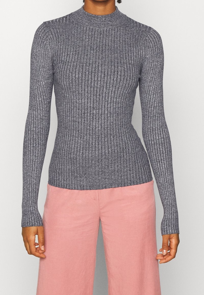 Femme portant un haut gris côtelé à manches longues ajusté et un pantalon rose ample taille haute, debout devant un fond clair uni.