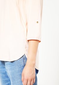Blouse rose clair à manches trois-quarts, avec un col rond et un petit bouton doré près du poignet. Associée à un jean bleu.