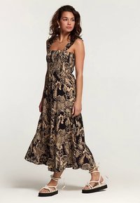 Robe longue avec un motif floral noir et beige, bretelles ajustables et jupe fluide. Portée avec des sandales plates et des accessoires minimalistes.