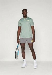 Maglietta polo verde chiaro con colletto, pantaloni corti grigi, scarpe da ginnastica bianche; tiene in mano una racchetta da tennis e una pallina. Abbigliamento sportivo casual con dettagli minimi.