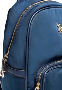 Mörkblå nylonryggsäck med gyllene dragkedjor, med en slät textur och en liten dekorativ logotyp på framlappen.