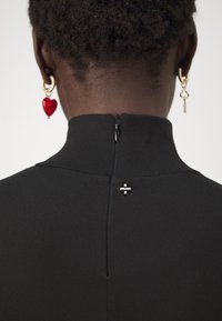 Zwarte jurk met een hoge halslijn, met een ritssluiting op de rug en een zilveren accent bij de hals. Het model draagt rode hartvormige oorbellen.
