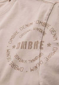 Béžové bavlněné tričko s kulatým béžovým potiskem a textem "OMBRE DENIM" a "ORIGINAL VINTAGE" v různých písmenech a orientacích.