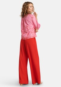 Rosa blumenmuster Bluse mit lockerer Passform, kombiniert mit weiten roten Hosen aus strukturiertem Stoff. Minimale Metallverzierungen.