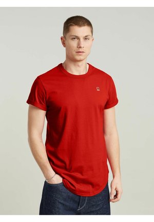 Homme portant un t-shirt rouge à manches courtes et un jean bleu foncé, debout avec une main dans la poche devant un fond clair uni.