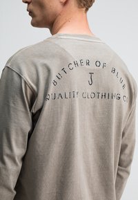 Langärmliges, hellgraues Baumwollhemd mit Rundhalsausschnitt und bedrucktem Text auf der Rückseite, darunter "BUTCHER OF BLUE" und "QUALITY CLOTHING CO."