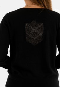 Un pull noir avec un emblème texturé en argent au dos. Le tissu est lisse, avec un col rond classique et des manches longues.