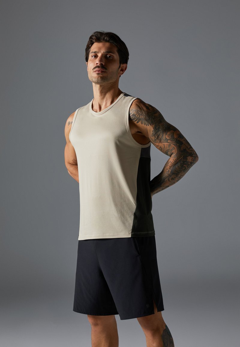 Ærmeløs tanktop i beige med en sort sidepanel; lavet af glat stof. Båret med mørke shorts, der viser en muskuløs bygning og tatoveringer.