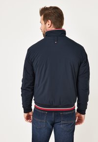 Mann mit braunen Haaren und Bart, der eine marineblaue Jacke mit einem rot-weißen gestreiften Kragen und Saum trägt, kombiniert mit einer dunkelblauen Jeans, von hinten blickend.
