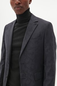 Blazer azul marino con un sutil patrón de camuflaje tonal, diseño de una sola botonadura, solapas con muesca y un bolsillo en el pecho, combinado con un cuello de tortuga negro.