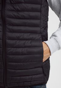 Schwarz gepolsterte Weste mit horizontalen Steppnähten, Reißverschluss und Seitentaschen. Der Träger trägt darunter einen hellgrauen Sweatshirt.