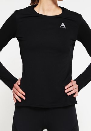 T-shirt de sport noir à manches longues en tissu léger et doux. Il présente un col rond et un petit logo argenté sur le côté gauche en haut.