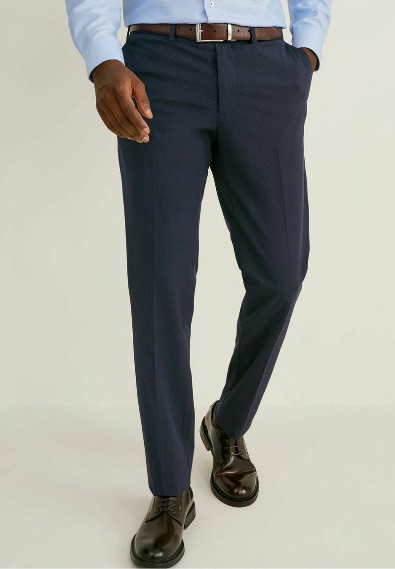 Homme portant un pantalon de costume bleu marine, une chemise bleu clair, une ceinture en cuir marron et des chaussures de ville marron foncé cirées marchant en avant.