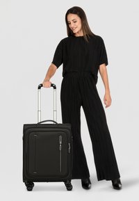Valigia nera con ruote, superficie testurizzata, due tasche frontali e manico telescopico, posizionata accanto a una persona in un abito plissettato.