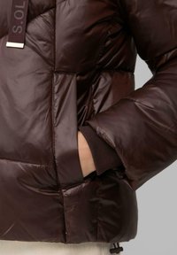 Braune Steppjacke mit glänzender, strukturierter Oberfläche. Verfügt über eine Reißverschlusstasche, gerippte Bündchen und einen markierten Riemen auf der Vorderseite.
