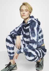 Sweatshirt com capuz em tie-dye e calças combinando em azul e branco, com uma textura suave e um corte descontraído, acompanhados de sapatilhas de atletismo cinzas.