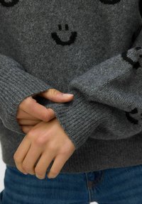 Szary sweter wykonany z miękkiego materiału dzianinowego, z prążkowanymi rękawami i wzorami uśmiechniętej buźki w kolorze czarnym. Ręce złożone na mankietach.