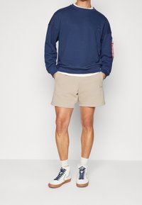 Felpa blu navy con scollo a girocollo, vestibilità rilassata e tasca laterale; pantaloni corti beige; scarpe alte bianche con lacci blu e calzini chiari.