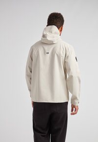 Lys beige vandtæt jakke med hætte, rygsøm detaljer, elastiske manchetter og et logo på venstre ærme. Glat tekstur.