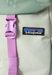 Mochila Patagonia com um corpo cinza claro, detalhes em verde menta, uma fivela roxa e um patch de logo preto com gráficos de montanhas.