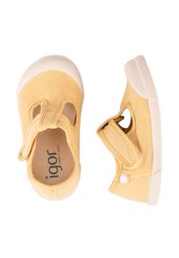 Zapatillas de lona amarillas con punta redondeada, correa de Velcro y detalles laterales recortados. La suela es de goma blanca con textura antideslizante.