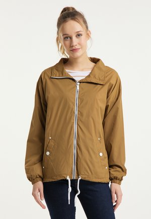 Jeune femme aux cheveux clairs attachés en queue de cheval, portant une veste marron à zip et un jean foncé, debout devant un fond clair uni.