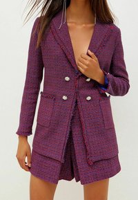Blazer croisé en tweed violet avec une finition texturée, dotée de deux poches avant et de boutons embellis. Des bords ourlés ajoutent des détails.