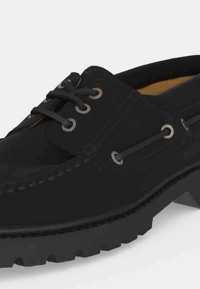 Μαύρο δερμάτινο παπούτσι τύπου boat shoe με χοντρή λαστιχένια σόλα, ασημένιες ρέγγες και μαύρα κορδόνια, εμφανιζόμενο σε κοντινή γωνιακή λήψη.
