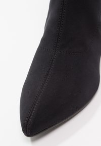 Botte noire à bout pointu avec des coutures visibles sur un tissu lisse, montrée de face légèrement inclinée sur le côté sur fond blanc.