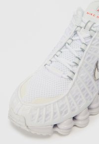 Sapata atlética branca com parte superior em malha, sobreposição estruturada, sola arredondada proeminente e acabamentos prateados. Apresenta atacadores e marcação na língua.