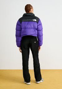 Puffer-jakke i lilla og svart, cropped design, skinnende materiale med horisontal quiltet tekstur. Kombinert med svarte kordfløyelsbukser.
