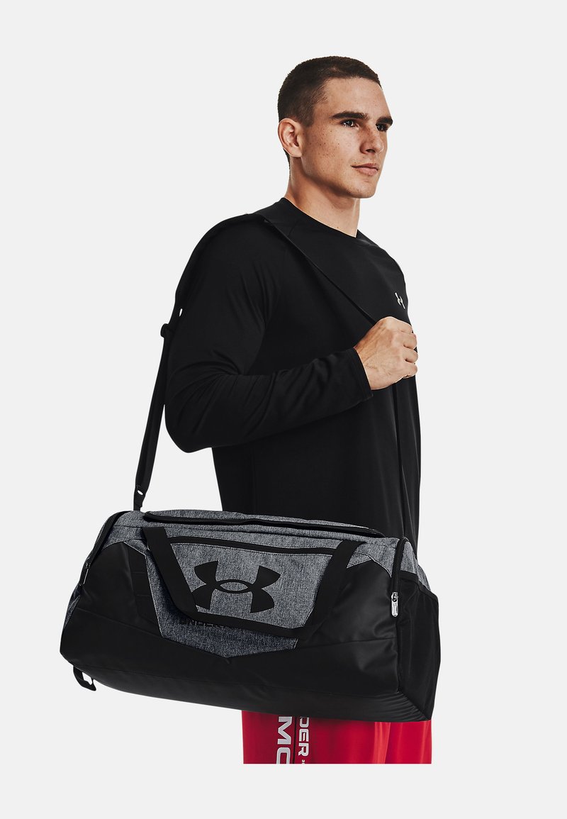 Mann trägt schwarzes Langarmshirt und rote Shorts und trägt eine große schwarze und graue Under Armour Sporttasche über der Schulter.