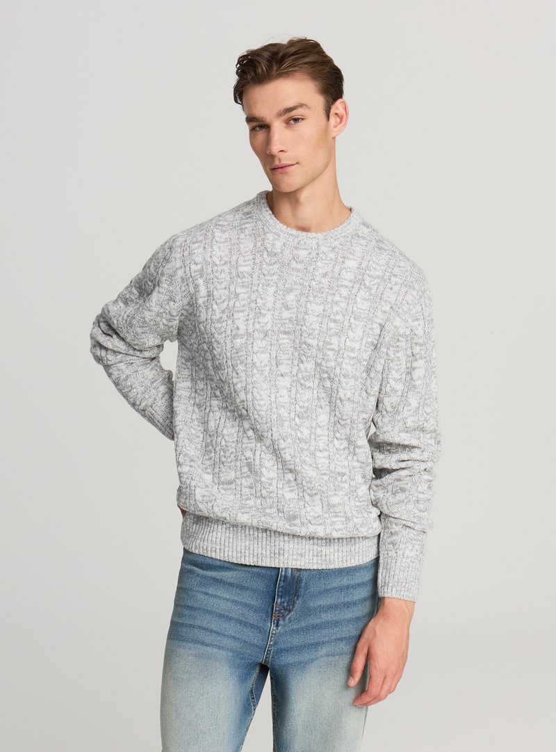 Szary sweter dzianinowy z fakturą w warkoczowy wzór, okrągłym dekoltem i prążkowanymi ściągaczami, zestawiony z jasnoniebieskimi dżinsami.