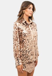 Top con paillettes in oro rosa, maniche lunghe, colletto con bottoni e una vestibilità rilassata. Rivestito da una superficie interamente decorata con paillettes rotonde che creano un effetto scintillante.