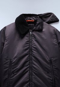 Černá prošívaná bunda s texturovaným, falešným beránkovým límcem, zipem a dvěma bočními kapsami. Hladká, polstrovaná látka s oranžovým akcentem.