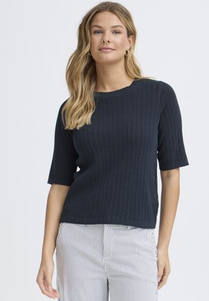 Jeune femme aux longs cheveux blonds portant des boucles d'oreilles anneaux dorées, un haut en tricot noir à manches courtes, et un pantalon blanc à fines rayures verticales.