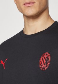 T-shirt nera in cotone con logo Puma rosso ed emblema del AC Milan rosso. Design con collo rotondo, maniche corte e texture liscia.