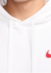 Vit huvtröja med dragsnören och en röd broderad logotyp på bröstet. Tyget verkar mjukt med en klassisk, avslappnad passform.