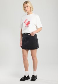 Una camiseta blanca con un gráfico de cereza roja y texto, combinada con una falda de mezclilla negra. Unos mocasines negros brillantes y calcetines blancos completan el look.