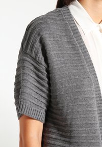 Grå stickad cardigan med ribbade kortärmar och ett strukturerat vertikalt mönster. Har en V-halsdesign och kontrasterar mot en vit skjorta.