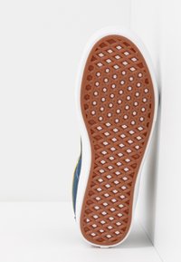 Podešev boty s texturovanou hnědou gumou a vzorem ve tvaru diamantu, lemovaná bílým okrajem. Na bocích jsou viditelné modré a žluté akcenty.