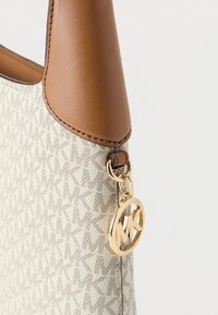 Beżowa i brązowa designerska torebka z powtarzającym się wzorem logo "MK" i złotą okrągłą zawieszką z logo "MK" przymocowaną do uchwytu.