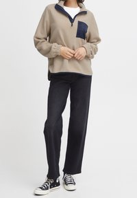 Person iført beige fleece-pullover med marineblå lomme, sorte straight-leg jeans og sorte høj-top sneakers med hvide snørebånd.
