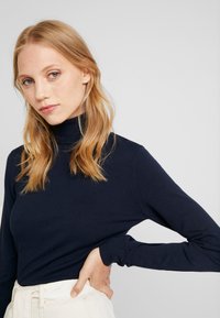 Marinblå turtlenecktröja i stickad kvalitet, figurnära snitt, långa ärmar, ribbad krage och manschetter, bärs med ljusa, högmidjade byxor.