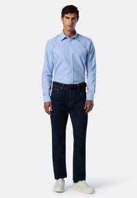 Camicia azzurro chiaro con bottoni, jeans blu scuro con cintura e sneaker bianche. La camicia presenta un colletto e un logo discreto.