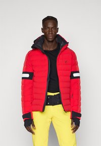 Rote Pufferjacke mit Reißverschluss, schwarzen Akzenten und einem weißen Streifen am Ärmel. Kombiniert mit gelben Hosen, die ein sportliches Design zeigen.