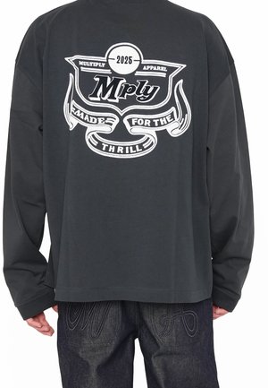 Maglietta nera a maniche lunghe con testo grafico bianco "Mply 2025 Multiply Apparel Made for the Thrill" sul retro, indossata con jeans scuri che mostrano tasche ricamate.