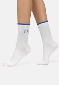 Hvide bomuldsokker med lodret ribbede detaljer, blå kant foroven og et blåt smilende ansigt grafisk tryk på siden. Kort design, fodlængden synlig.