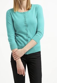 Ljus turkos kofta med rund halsringning, knäppning fram med knappar och trekvartslånga ärmar. Slät stickad textur, smal passform.