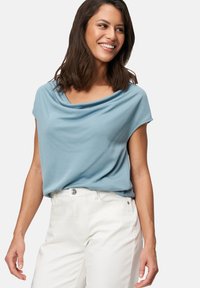 Light blue short-sleeve top with a draped neckline, made of soft fabric, paired with white high-waisted pants. Smooth texture, relaxed fit.
